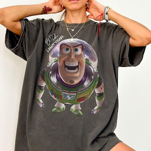May include: A charcoal gray oversized t-shirt featuring a large graphic of the character Buzz Lightyear from Toy Story. The character is depicted in a clear helmet with green, purple, and white armor. The text BUZZ LIGHTYEAR is printed above.
