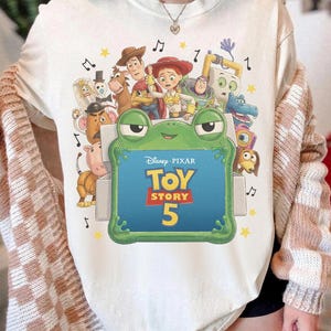 Może przedstawiać: Kremowy t-shirt z nadrukiem graficznym przedstawiającym postacie z filmu Disney Pixar Toy Story, w tym Chudego, Jessie i Buzza Astrala. Duża zielona ramka w kształcie żaby otacza napis Disney Pixar Toy Story 5 z ż&oacute;łtymi gwiazdkami.