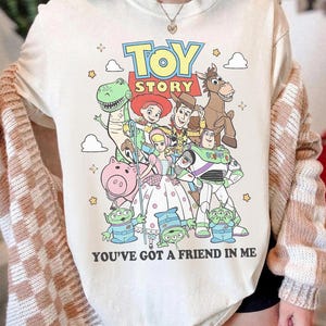 May include: A cream-colored t-shirt featuring a colorful graphic of Toy Story characters including Woody, Buzz Lightyear, Jessie, Rex, Bullseye, Hamm, Bo Peep, and the Aliens. The text reads TOY STORY and YOU'VE GOT A FRIEND IN ME with small stars.