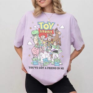 May include: A light purple short-sleeve t-shirt featuring a colorful graphic print of Toy Story characters including Woody, Buzz Lightyear, Jessie, Rex, Bullseye, Hamm, Bo Peep, and the Aliens, with the text Toy Story and You've Got A Friend In Me.