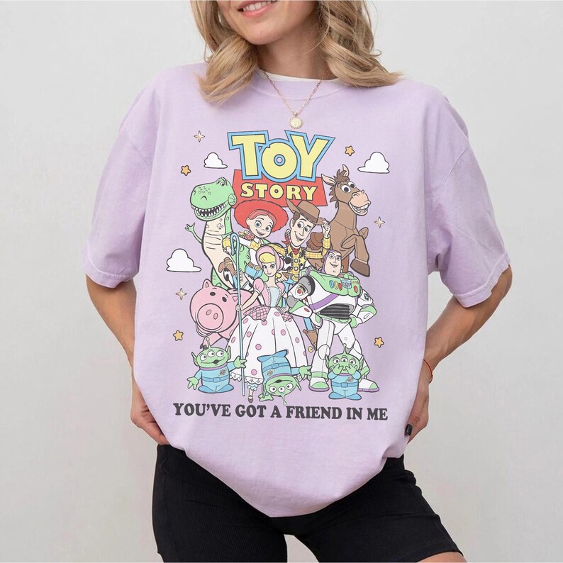 May include: A light purple short-sleeve t-shirt featuring a colorful graphic print of Toy Story characters including Woody, Buzz Lightyear, Jessie, Rex, Bullseye, Hamm, Bo Peep, and the Aliens, with the text Toy Story and You've Got A Friend In Me.