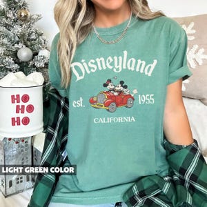 K&ouml;nnte beinhalten: Hellgr&uuml;nes T-Shirt mit dem Wort "Disneyland" in wei&szlig;er Schrift. Das Shirt zeigt eine Grafik von Mickey und Minnie Mouse in einem roten Auto mit dem Text "est. 1955 CALIFORNIA". Das Shirt ist ein bequemer, l&auml;ssiger Stil.