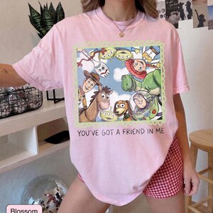 May include: A light pink t-shirt featuring a colorful graphic of characters from the movie Toy Story, including Woody, Buzz Lightyear, and Rex. The text "YOU'VE GOT A FRIEND IN ME" is printed below the graphic. The shirt is made of a soft material.
