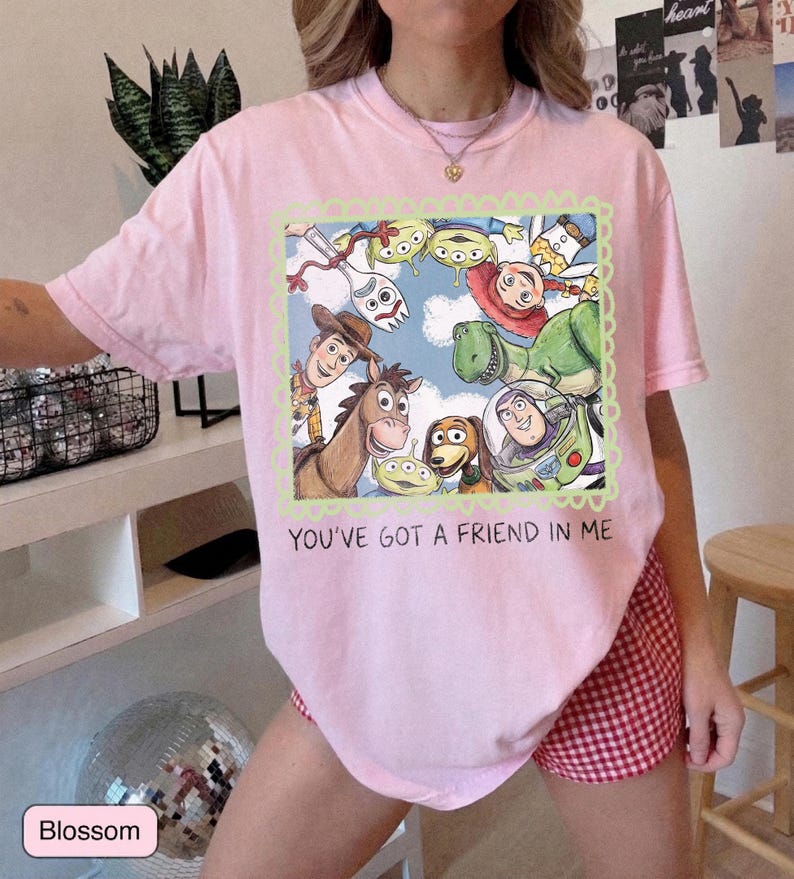 May include: A light pink t-shirt featuring a colorful graphic of characters from the movie Toy Story, including Woody, Buzz Lightyear, and Rex. The text "YOU'VE GOT A FRIEND IN ME" is printed below the graphic. The shirt is made of a soft material.