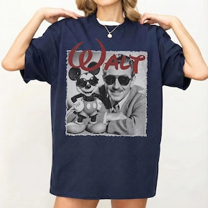 May include: Navy blue t-shirt featuring a black and white image of Walt Disney with Mickey Mouse wearing sunglasses. The word "WALT" is printed in red above the image. The shirt is a classic crew neck style.