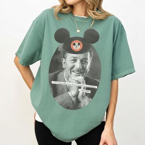 May include: Green t-shirt featuring a black and white image of Walt Disney wearing a Mickey Mouse hat. The shirt includes the quote, "I love Mickey Mouse more than any woman I have ever known."
