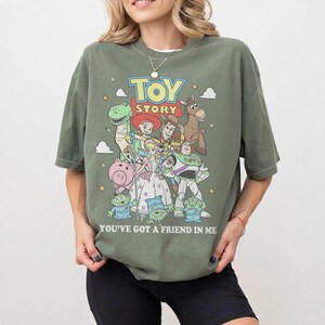 May include: An olive green t-shirt featuring a graphic print of Toy Story characters including Woody, Buzz Lightyear, Jessie, Rex, Hamm, Bullseye, and the Aliens. The text Toy Story and You've Got A Friend In Me is printed in white and yellow.