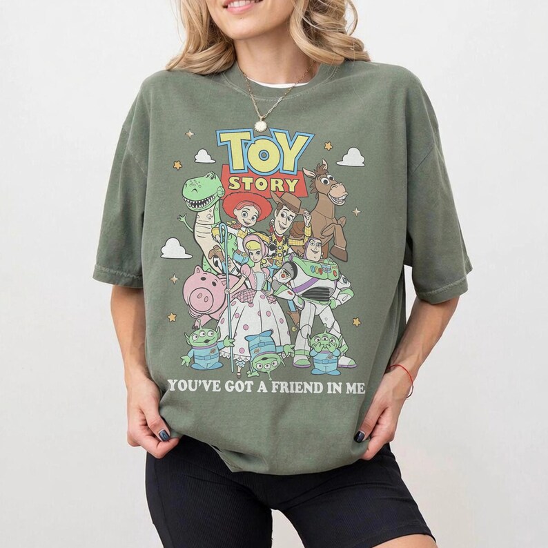 May include: An olive green t-shirt featuring a graphic print of Toy Story characters including Woody, Buzz Lightyear, Jessie, Rex, Hamm, Bullseye, and the Aliens. The text Toy Story and You've Got A Friend In Me is printed in white and yellow.