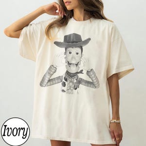 May include: An ivory-colored short-sleeve t-shirt featuring a black and white graphic print of the character Woody from Toy Story. The character is depicted wearing a cowboy hat, a neckerchief, and a vest, with hands raised near the face.