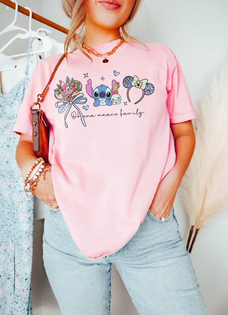 Puede incluir: Camiseta rosa claro con un dise&ntilde;o de Stitch, Angel y orejas de Mickey Mouse florales. La frase "Ohana means family" est&aacute; impresa en negro. La camiseta es de un material suave.