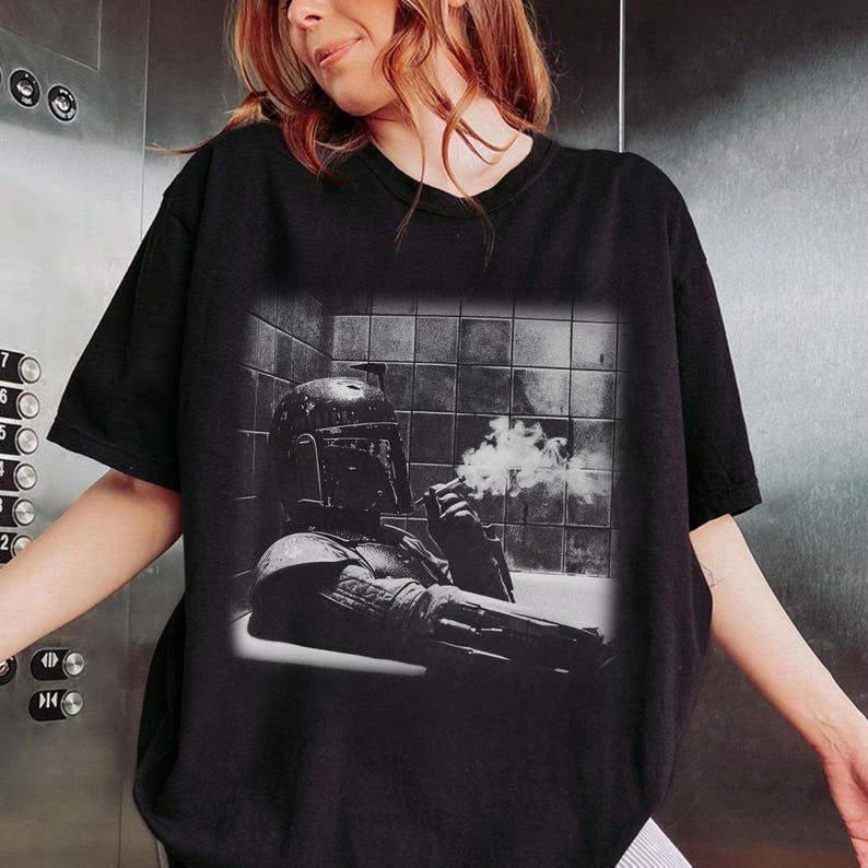 May include: A black short-sleeve t-shirt featuring a monochromatic graphic print of a helmeted character in a tiled room, holding a smoking object. The design shows the character from the chest up, set against a background of square wall tiles.