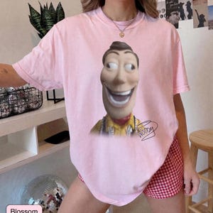 May include: A light pink short-sleeve t-shirt featuring a large graphic of the character Woody from Toy Story with a wide, exaggerated smile. Below the character, the name Woody is written in a black cursive script. The shirt has a crew neckline.