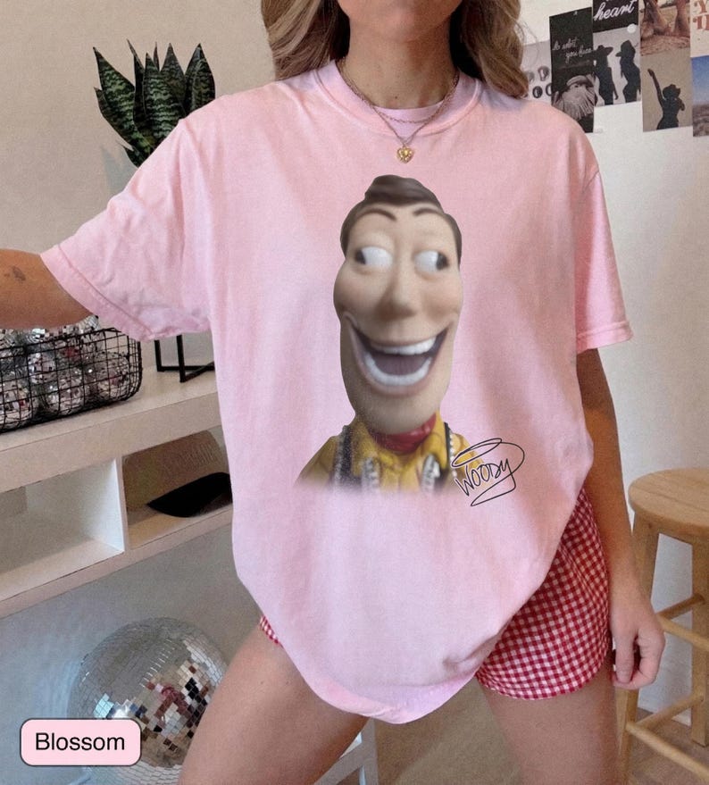 May include: A light pink short-sleeve t-shirt featuring a large graphic of the character Woody from Toy Story with a wide, exaggerated smile. Below the character, the name Woody is written in a black cursive script. The shirt has a crew neckline.