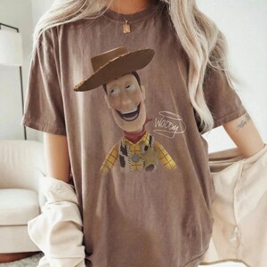 May include: A brown short-sleeved t-shirt featuring a graphic print of the character Woody from Toy Story wearing a cowboy hat, yellow plaid shirt, and sheriff badge, with the name Woody written in white cursive script next to the character.