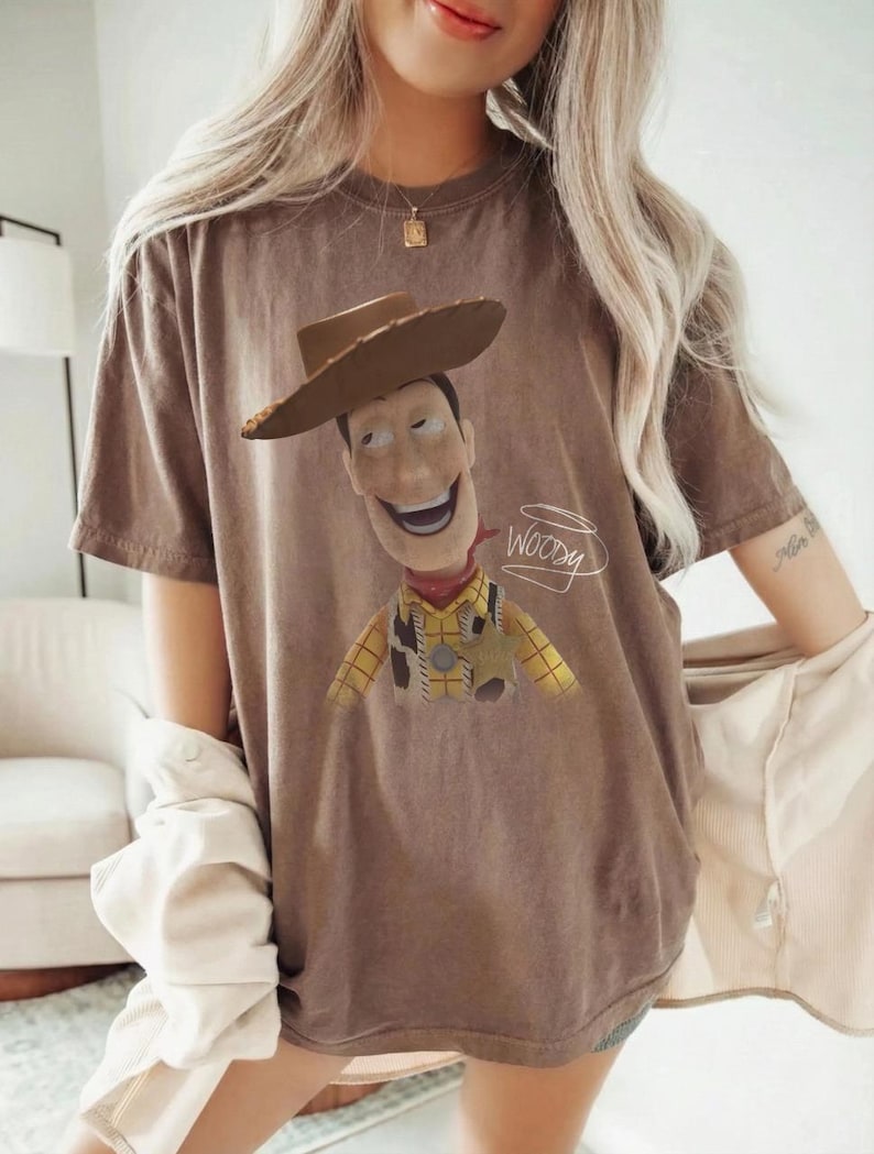 May include: A brown short-sleeved t-shirt featuring a graphic print of the character Woody from Toy Story wearing a cowboy hat, yellow plaid shirt, and sheriff badge, with the name Woody written in white cursive script next to the character.