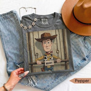 May include: A charcoal gray t-shirt featuring a graphic of a cowboy character wearing a brown hat, yellow shirt, and cow-print vest. The character holds a sign that reads "WOODY". The shirt is paired with blue denim jeans, a silver flower-shaped necklace, and a tan hat.