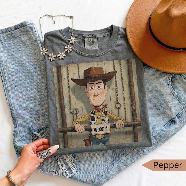 May include: A charcoal gray t-shirt featuring a graphic of a cowboy character wearing a brown hat, yellow shirt, and cow-print vest. The character holds a sign that reads "WOODY". The shirt is paired with blue denim jeans, a silver flower-shaped necklace, and a tan hat.