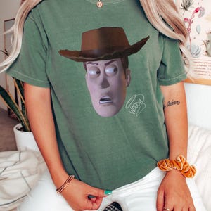 May include: A sage green short-sleeve t-shirt featuring a graphic print of the character Woody from Toy Story wearing a brown cowboy hat. The character has a surprised expression with wide eyes and an open mouth. The word Woody is written in cursive.