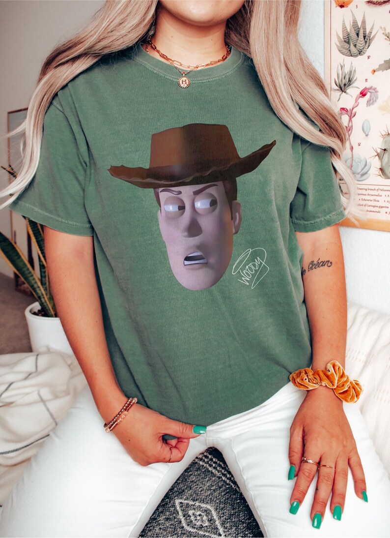 May include: A sage green short-sleeve t-shirt featuring a graphic print of the character Woody from Toy Story wearing a brown cowboy hat. The character has a surprised expression with wide eyes and an open mouth. The word Woody is written in cursive.
