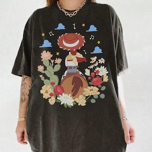 May include: A black short-sleeve t-shirt featuring a graphic print of a cowgirl figure on a horse, surrounded by cacti, various flowers, blue clouds, yellow stars, and musical notes. The design is centered on the front of the garment.