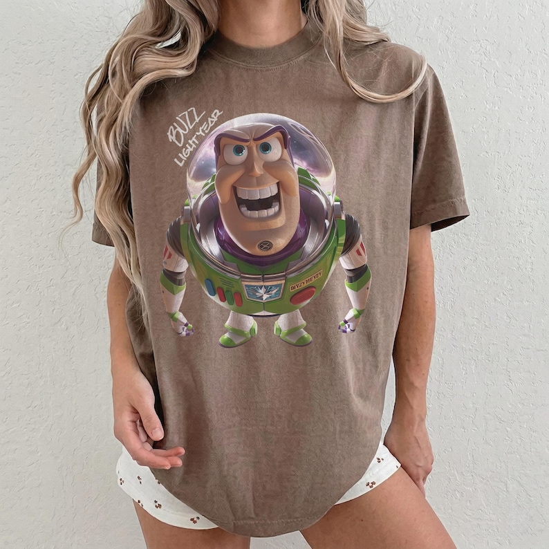 May include: A brown short-sleeve t-shirt featuring a graphic of the character Buzz Lightyear from Toy Story. The character is depicted in a clear helmet with green, white, and purple armor. The text BUZZ LIGHTYEAR is printed in white above.