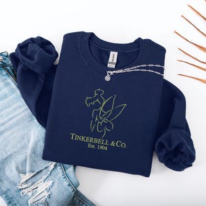 Pode incluir: Um moletom azul marinho com o contorno verde lim&atilde;o de uma fada e o texto "Tinkerbell & Co. Est. 1904." Um colar de prata com um pingente circular repousa sobre o moletom. Cal&ccedil;as de ganga azul claro s&atilde;o vis&iacute;veis no fundo.