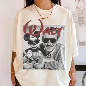 May include: Cream-colored t-shirt featuring a vintage graphic of Mickey Mouse wearing sunglasses next to a black and white image of Walt Disney. The word "WALT" is printed in red above the images.