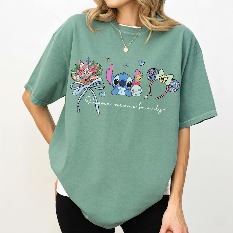 Puede incluir: Camiseta verde salvia con un gr&aacute;fico de Stitch, Angel y accesorios tem&aacute;ticos. El dise&ntilde;o incluye un lazo, detalles florales y orejas de rat&oacute;n. El texto "Ohana means family" est&aacute; escrito en cursiva debajo del gr&aacute;fico.