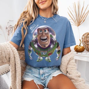 May include: A blue short-sleeve t-shirt featuring a graphic of the character Buzz Lightyear from Toy Story. The character is depicted in a space suit with green, purple, and white details. The text BUZZ LIGHTYEAR is printed in white above the character.