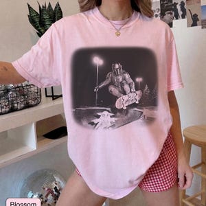 May include: A light pink short-sleeve t-shirt featuring a black and white graphic print of a character in armor skateboarding in a bowl, with a small creature watching from below. The graphic is set against a dark, blurred rectangular background.