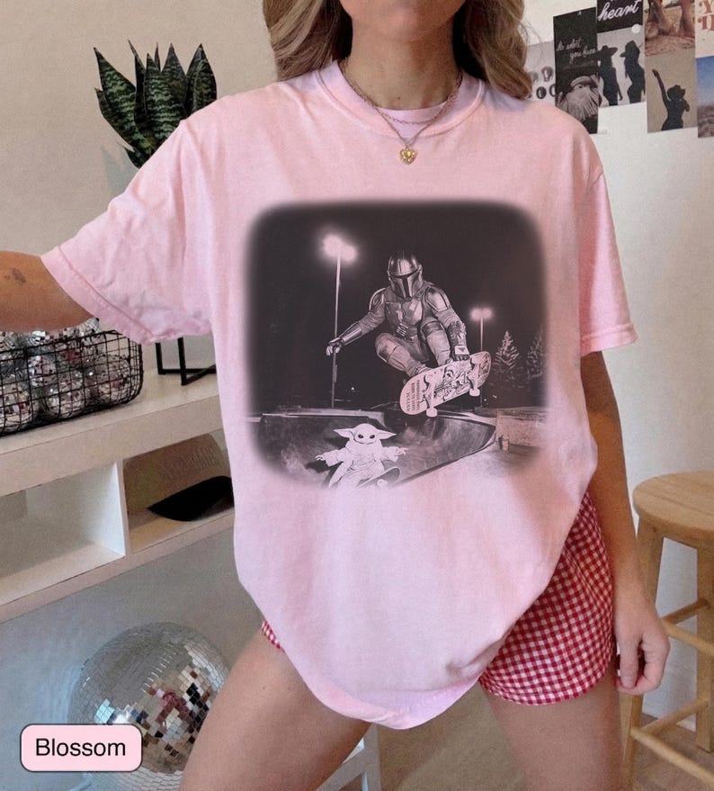 May include: A light pink short-sleeve t-shirt featuring a black and white graphic print of a character in armor skateboarding in a bowl, with a small creature watching from below. The graphic is set against a dark, blurred rectangular background.