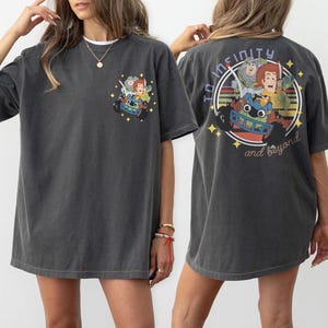 Op de afbeelding: Donkergrijs t-shirt met een kleurrijke afbeelding van stripfiguren in een raceauto, met de tekst "To Infinity and Beyond" op de achterkant. De voorkant van de shirt heeft een kleinere versie van de afbeelding. Een casual, comfortabel t-shirt.