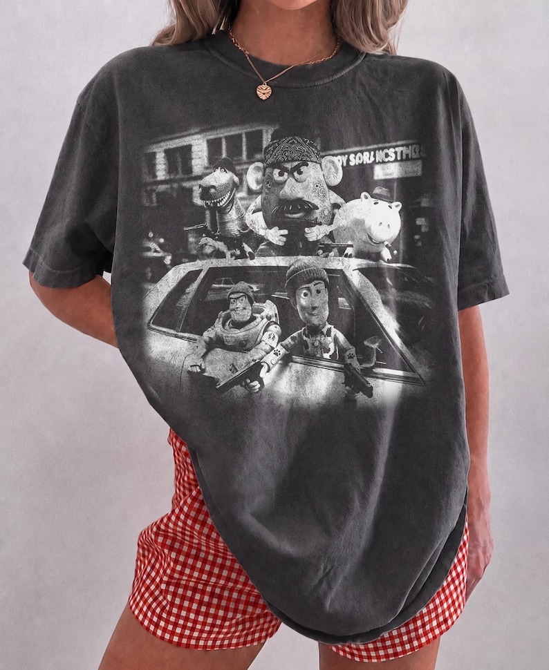May include: A charcoal gray short-sleeve t-shirt featuring a black and white graphic print of five animated characters in and on top of a car. The characters include a dinosaur, a potato figure, a pig, a space ranger, and a cowboy, all holding guns.