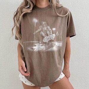 May include: A brown short-sleeve t-shirt featuring a white graphic print of a helmeted figure riding a skateboard, with a small creature standing nearby. The design includes stylized lighting elements and trees in a monochromatic, distressed style.