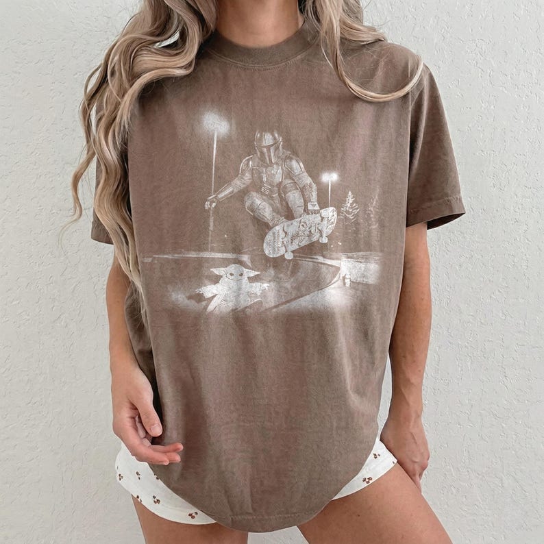 May include: A brown short-sleeve t-shirt featuring a white graphic print of a helmeted figure riding a skateboard, with a small creature standing nearby. The design includes stylized lighting elements and trees in a monochromatic, distressed style.
