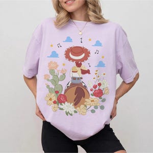 May include: A light purple t-shirt featuring a graphic print of a cowgirl character wearing a red hat, riding a brown horse. The design includes green cacti, red and yellow flowers, blue clouds, and black musical notes scattered around the central figure.