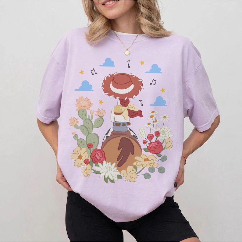 May include: A light purple t-shirt featuring a graphic print of a cowgirl character wearing a red hat, riding a brown horse. The design includes green cacti, red and yellow flowers, blue clouds, and black musical notes scattered around the central figure.
