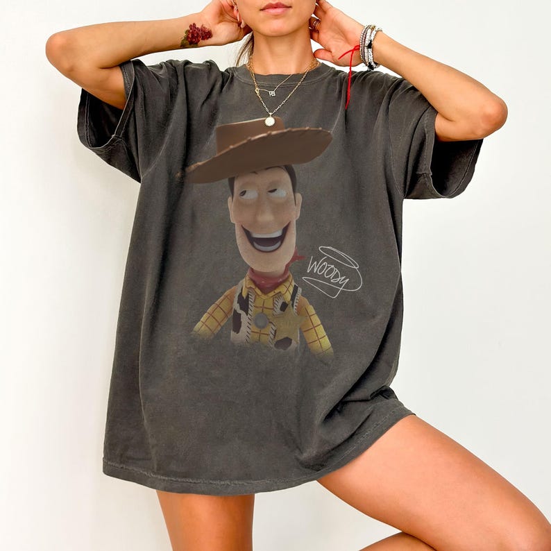 May include: A charcoal gray short-sleeve t-shirt featuring a graphic print of the character Woody from Toy Story wearing a brown cowboy hat. The design includes a yellow and red plaid shirt, a cow-print vest, and the handwritten text Woody in white.