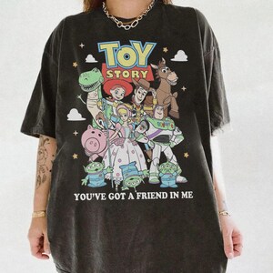 May include: A charcoal gray short-sleeve t-shirt featuring a colorful graphic print of Toy Story characters including Woody, Buzz Lightyear, Jessie, Rex, Hamm, and the Aliens. The text reads TOY STORY and YOU'VE GOT A FRIEND IN ME.