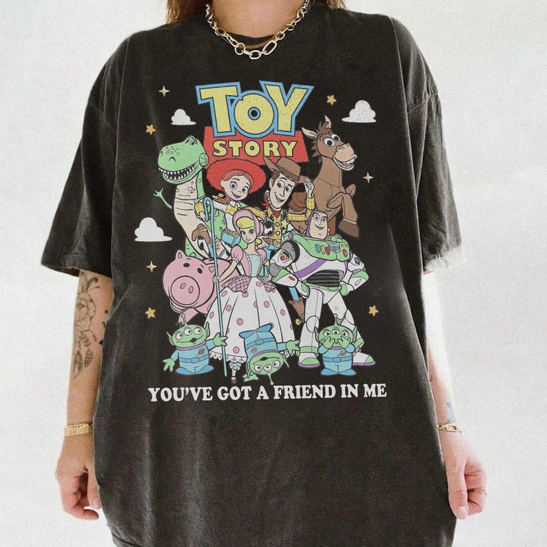 May include: A charcoal gray short-sleeve t-shirt featuring a colorful graphic print of Toy Story characters including Woody, Buzz Lightyear, Jessie, Rex, Hamm, and the Aliens. The text reads TOY STORY and YOU'VE GOT A FRIEND IN ME.