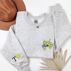 May include: A light gray crewneck sweatshirt featuring an embroidered green three-eyed alien character on the chest. The left sleeve displays an embroidered black letter T followed by a small green heart. The garment has a standard ribbed collar.