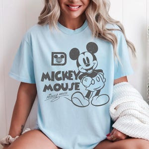 May include: Light blue t-shirt featuring a distressed graphic of Mickey Mouse. The design includes the text "Mickey Mouse" and a signature. The shirt has short sleeves and a crew neck.