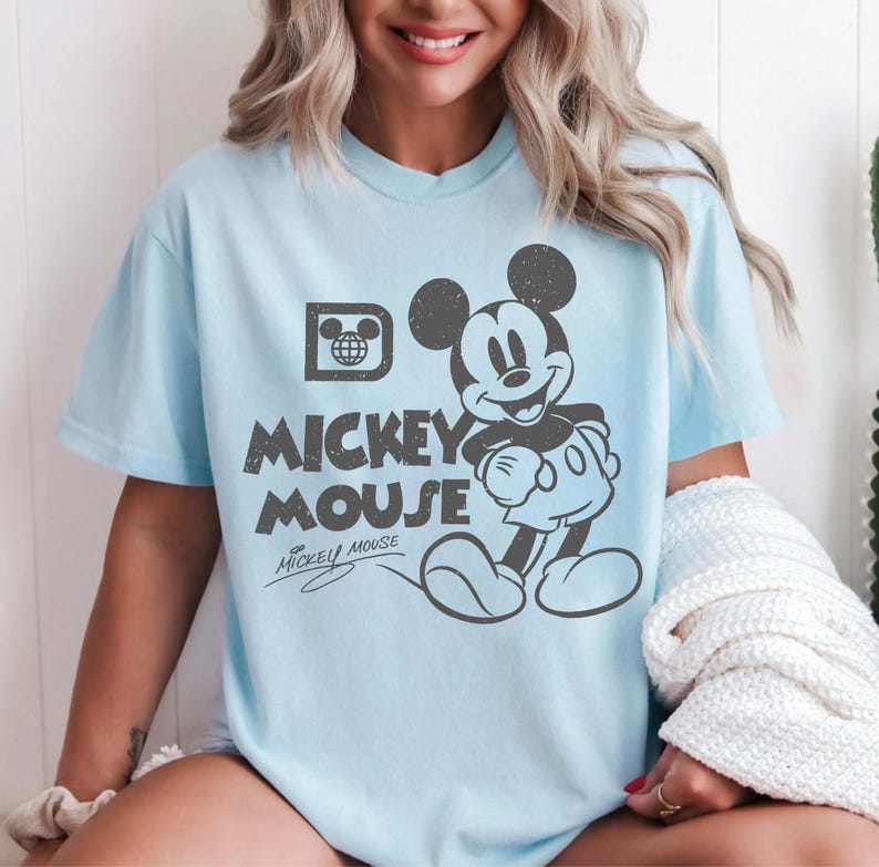 May include: Light blue t-shirt featuring a distressed graphic of Mickey Mouse. The design includes the text "Mickey Mouse" and a signature. The shirt has short sleeves and a crew neck.