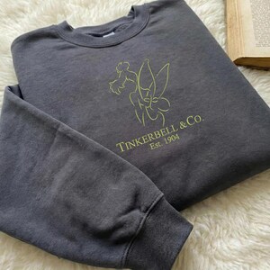 Pode incluir: Um moletom cinza carv&atilde;o com o texto "Tinkerbell & Co. Est. 1904" e um contorno verde lim&atilde;o de Tinkerbell. O moletom est&aacute; dobrado sobre uma superf&iacute;cie branca e fofa.