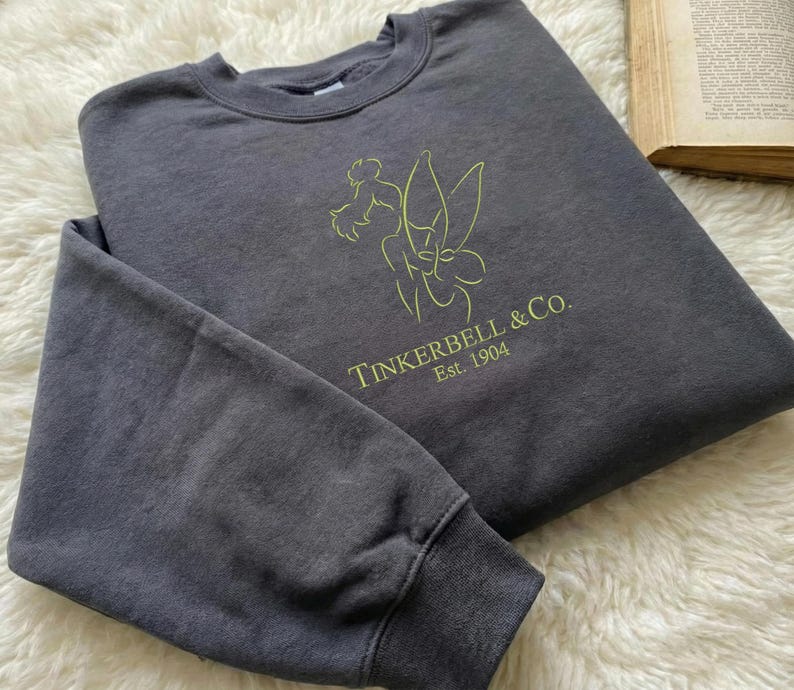 Pode incluir: Um moletom cinza carv&atilde;o com o texto "Tinkerbell & Co. Est. 1904" e um contorno verde lim&atilde;o de Tinkerbell. O moletom est&aacute; dobrado sobre uma superf&iacute;cie branca e fofa.