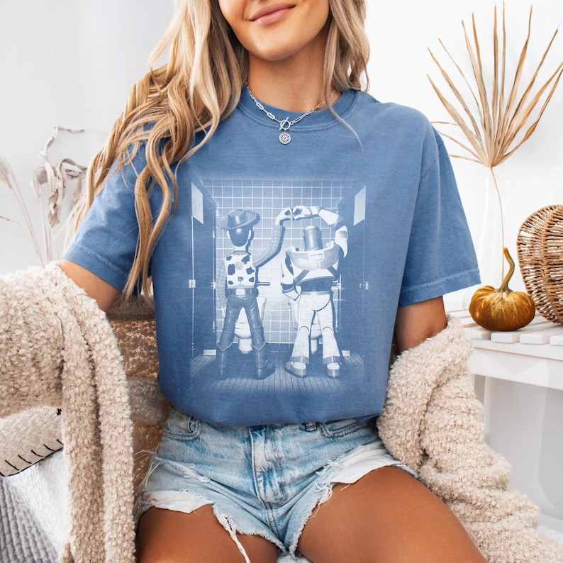 May include: A blue short-sleeve t-shirt featuring a white graphic print of two cartoon characters standing at urinals in a tiled restroom, with one character forming a heart shape with their hands above their head, set against a blue fabric background.