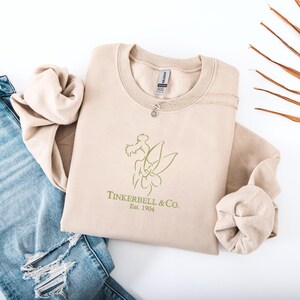 Pode incluir: Uma camisola bege com um contorno verde de uma fada e o texto "Tinkerbell & Co. Est. 1904." Um colar de prata com um pequeno pingente repousa sobre a camisola. A camisola est&aacute; dobrada ao lado de um par de jeans azuis.