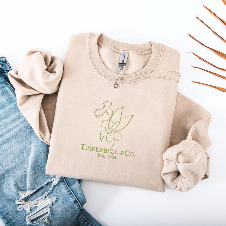 Pode incluir: Uma camisola bege com um contorno verde de uma fada e o texto "Tinkerbell & Co. Est. 1904." Um colar de prata com um pequeno pingente repousa sobre a camisola. A camisola est&aacute; dobrada ao lado de um par de jeans azuis.