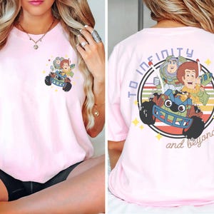 Op de afbeelding: Lichtroze T-shirt met een kleurrijke afbeelding van Toy Story-personages Woody, Buzz Lightyear en anderen in een speelgoedauto. De achterkant van het shirt heeft de tekst "To infinity and beyond" in een cirkel.