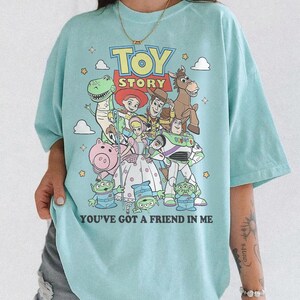 May include: A light blue t-shirt featuring a colorful graphic print of Toy Story characters including Woody, Buzz Lightyear, Jessie, Rex, Bullseye, Bo Peep, Hamm, and the Aliens, with the text TOY STORY and YOU'VE GOT A FRIEND IN ME printed on the front.